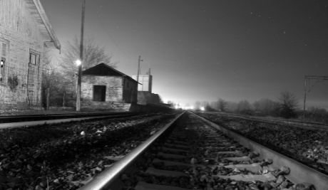A black and white landscape photograph of a railway track with buildings to the left hand side and trees along the horizon. 