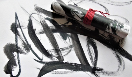 White paper with black painted brush marks in a calligraphy style. Top right is a scroll of paper, tied tightly with red string. The same black brush marks are on the outer scroll.