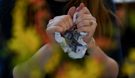 A close up of hands scrunching up a piece of artwork. In front, and out of focus, is a spray of bright yellow flowers.