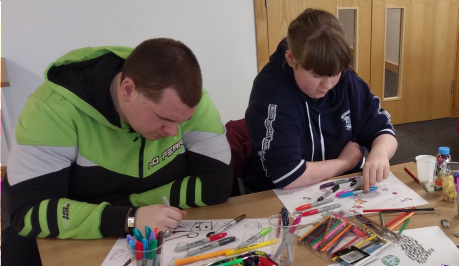 Two young people are concentrating on their work, on the table in front of them are pens and paper. The person on the left is drawing.