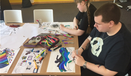 Two participants sit to the right of the picture, in front of them are pens, paper and their drawings. One person has drawn a figure swimming, the other person is drawing a picture of the world.