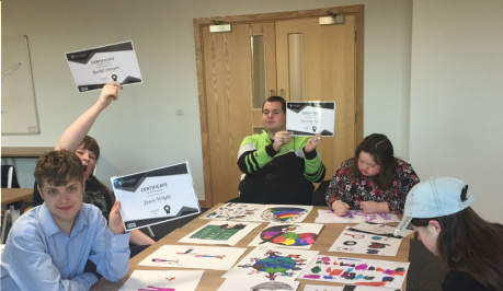 Five young people are sitting around a table, the table is covered in their drawings. Three of the young people are holding up their cerficates from Shropshire Council and DASH