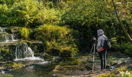 A photogrpah of a small waterfall surrounded by green vegetation. Facing the waterfall is a standing figure with a camera on a tripod. 