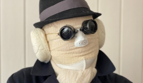 An image of a person with a fully covered head in bandages head, including ears. They wear a black ‘pork pie’ style hat, blacked out swimming goggles and a black mac style coat. Their white gloved hands are crossed in front of them just below their neck. 