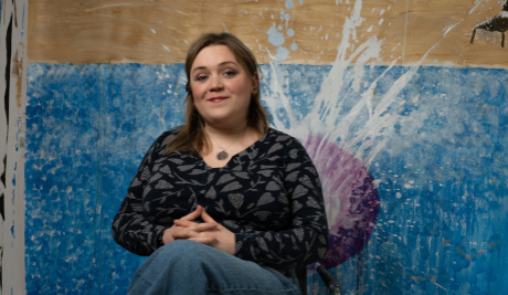 A portrait photograph showing a white woman with long brown hair sitting in a low swivel chair in front of a painting entitled Grace and John’s Biggest Splash.