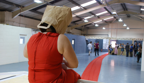 A woman in a red dress sits with her back to us in a large gallery space. She has a paper bag over her head and her red dress continues for the full length of the room right out of the entrance space, where there is a group of people.