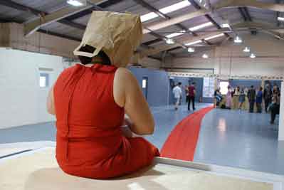 The artist Noemi Lakmaier sits on a wooden platform, she is wearing a red dress, which is so long it flows through the picture and out of the door.  She is wearing a paper bag on her head. In the background people are standing and talking. 