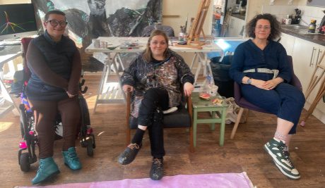 Artist Tanya Raabe Webber (left) and DASH Artistic Director/CEO Heather Peak (right) visit Grace (centre) in her studio. They sit in three chairs in a row.
