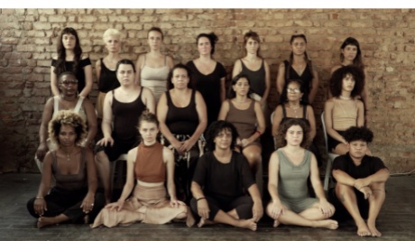 a group photograph of eighteen people (who appear to be women) arranged in three rows, the back row standing, the middle row seated on chairs and the front row seated on the floor. The subjects, all various ethnicities, are wearing muted coloured clothing