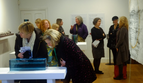 A photograph of visitors at Aidan's Llantarnam Grange Arts Centre exhibition. Two women lean over a light blue glass sculpture which one looks down at while the other looks to the left. Behind them, visitors look at the artwork or are in conversation.