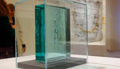 On a white plinth, is a perspex cube inside is a sea green coloured perspex rectangular box, inside which is held an image of a see through person, in the background are two women looking at the displays and a canvas painting of an orange, yellow,circle.