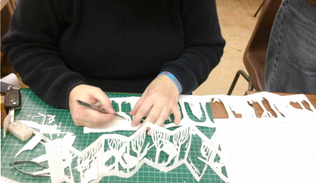 a person in a black jumper, carefully cutting white paper with an scalpel, into a intricate zig zagging folding book.
