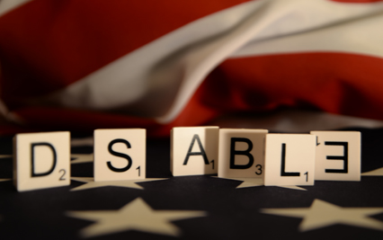 The shot is parallel to the standing Scrabble tiles/letters. The tiles read DSABLE from left to right of the image across the entire frame. The tiles are a quarter of the way up the page and it is a landscape shot. The first letter on the left is a black letter D (worth 2 points) a space is between the two tiles and then it is a letter S (worth 1 point) again a space nearly the size of a Scrabble tile but just slightly less. The Scrabble tile letter third across is an A with a shadow on the right side made by the tile in front of this tile the value is the frame (worth 1 point). The fourth tile across is B (worth 3 points). The fifth letter across is L (worth 1 point) and the final tile with letter E is upside down and in reverse. The tiles all stand on the blue fabric with white stars in the foreground. Behind the tiles is the red and white stripes of the fabric flag lifting away from the flat surface and layered folds of fabric concealing the striped pattern all slightly out of focus and blurred. The word is highlighting the fact disabled people are disabled by the frameworks  around them and not by their impairment. The E in reverse is a comment on 'the disable' who are still not quite allowed total inclusion into the every day. The letters signifying terminology around Dyslexia and children being labelled and left or forced to fit into a framework that doesn't work. The framework often created by adults to dis-empower disabled children.