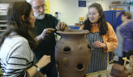 Three people, two young females with long brown hair and one older male with short grey hair and beard, are in the forefront of the photo, around a large garden pot being created.