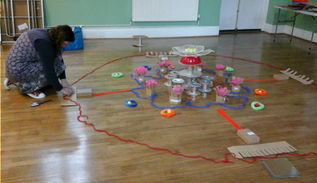 A dark-haired woman is kneeling down to the left of the photo, on a wooden floor, adjusting a piece of her mandala work laid out on the floor, made up of a range of objects made of plastic and paper in bright orange and deep blue and clear.