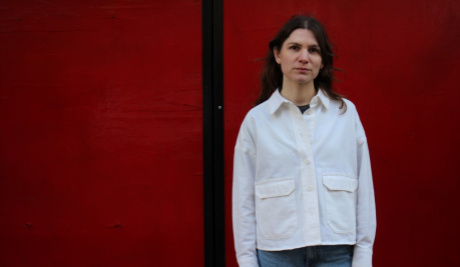 Hannah Wallis stands to the right hand side of the image against a dark red background. She wears a white shirt and blue jeans and faces towards the camera. 