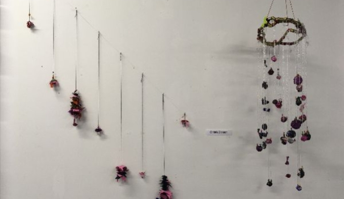 A photograph of eight pieces of string hanging vertically with coloured material at the end spaced in a descending diagonal line. To the right is mobile with pink and purple material attached to clear string, exhibited against a white background.   