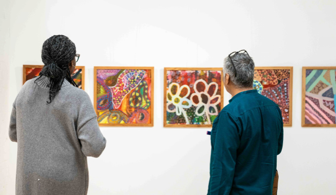Two people stand side by side in an art gallery, facing a wall of colourful, abstract paintings in wooden frames. They appear to be discussing or reflecting on the artworks together.