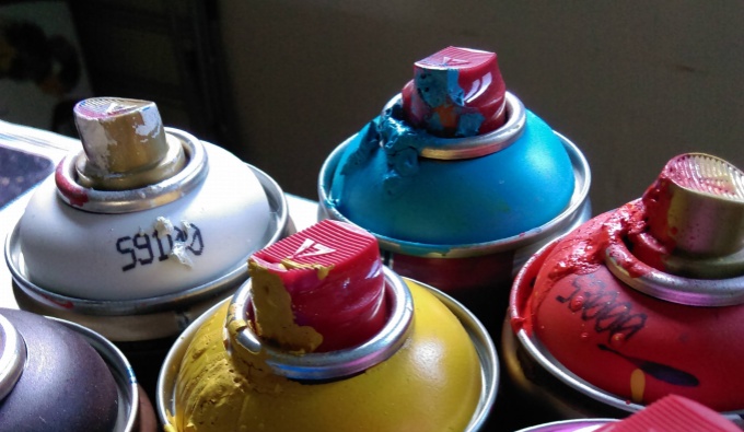 A view across the top if a group of spray cans against a dark background. Including a white, yellow, blue and red can. They have been used and paint dribbles from the nozzles.