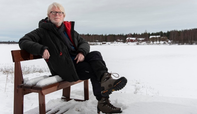 Aidan Moesby sits on a bench in a snowy landscape. He wears a dark coat, trousers and hiking boots. He has white blonde hair and rimless glasses.