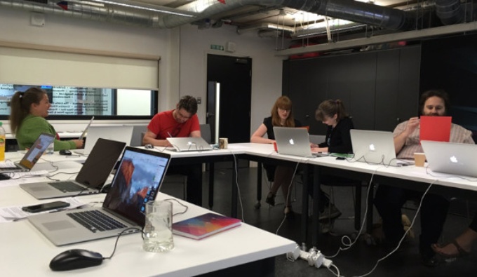 A meeting room with white desks arranged in a horseshoe shape. There are laptops on several of the desks and people looking at the screens.