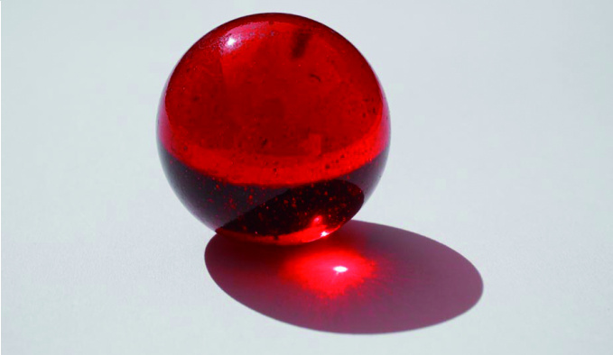 As single red glass marble on a grey background. The marble is lit from behind creating a red shadow, in the centre of which is a white spot