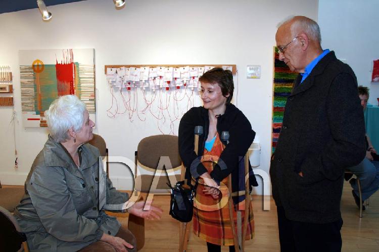 DASh Visual Arts Manager, Tanya Raabe is chatting to two visitors at the Artists Development Team Exhibition.  Tanya is standing in the centre of the image and is using crutches.  One visitor sits on the left, the other visitor is standing to the right.  On the white walls behind artworks are displayed.  Photograph Paula Dower copyright DASh.