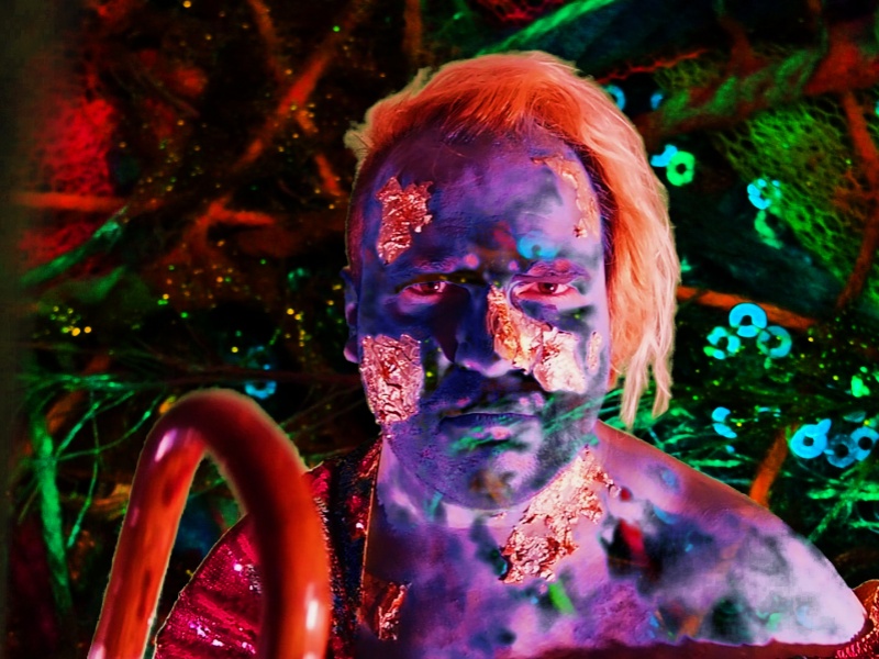Alex has a painted face and shoulders in blue and green. The top half of their red cane is just visible.