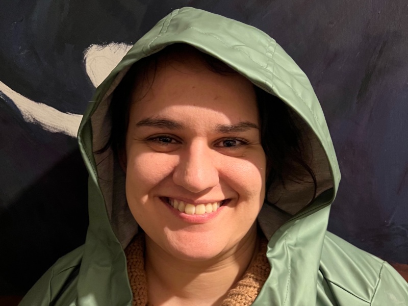 Anahita smiles at the camera. She wears a sage green rain mac with the hood pulled loosely over her head, with a sand coloured jumper underneath.