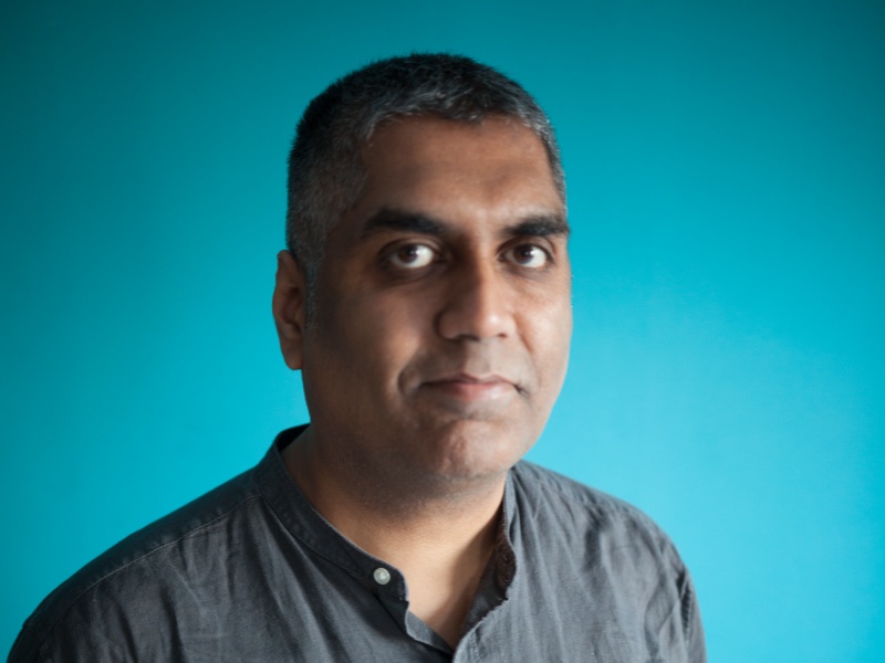 Ashok stands facing the camera against a turquoise background. He has short hair and wears a grey shirt. He looks directly at the camera.