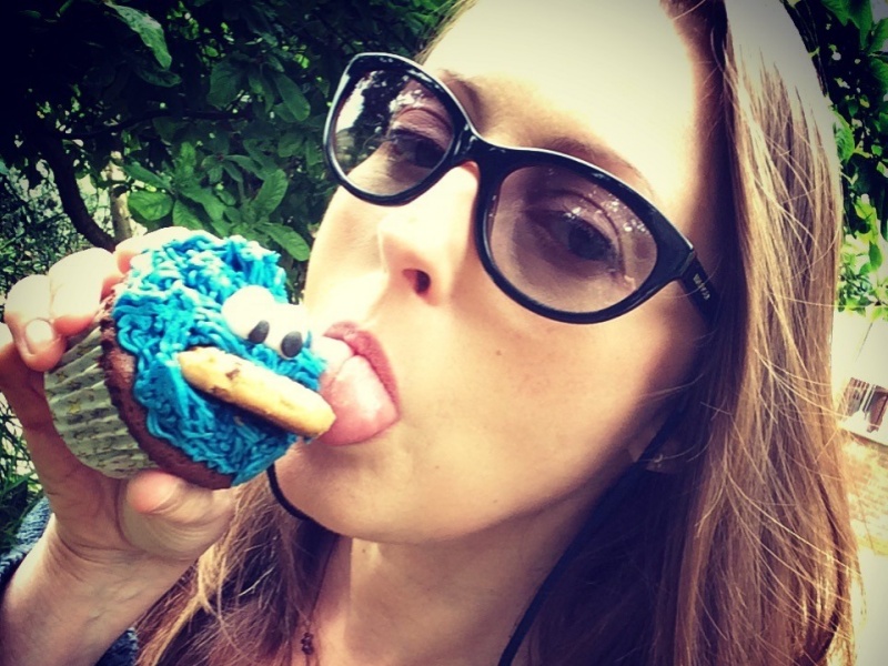 A close up portrait image of Caroline's face. She has long brown hair and wears sun glasses. Her head is tilted to the side as she looks at the camera and takes a bite from a cupcake with turquoise blue icing.