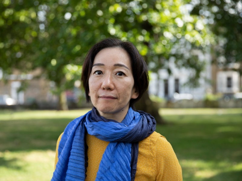 Artist Chisato Minamimura standing in a green space outdoors. She wears a mustard jumper and a two tone light and dark blue scarf.