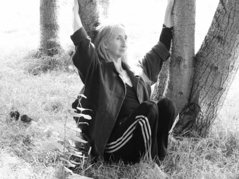 A black and white photograph of Christina sat on grass amongst young trees. She wears black tracksuit bottoms with 3 white stripes running up the side with a dark coloured jacket. She has her arms upstretched and holds the trunks of two trees.