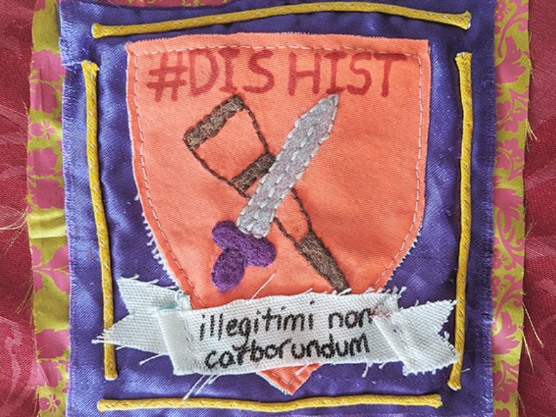 A square of yellow and pink fabric with a second smaller square of purple fabric attached on top. Sewn to this is a shield shape in orange cloth. Embroidered onto which is a crutch and a sword. Above in pen #DisHist and below illegitemi non carborundum.