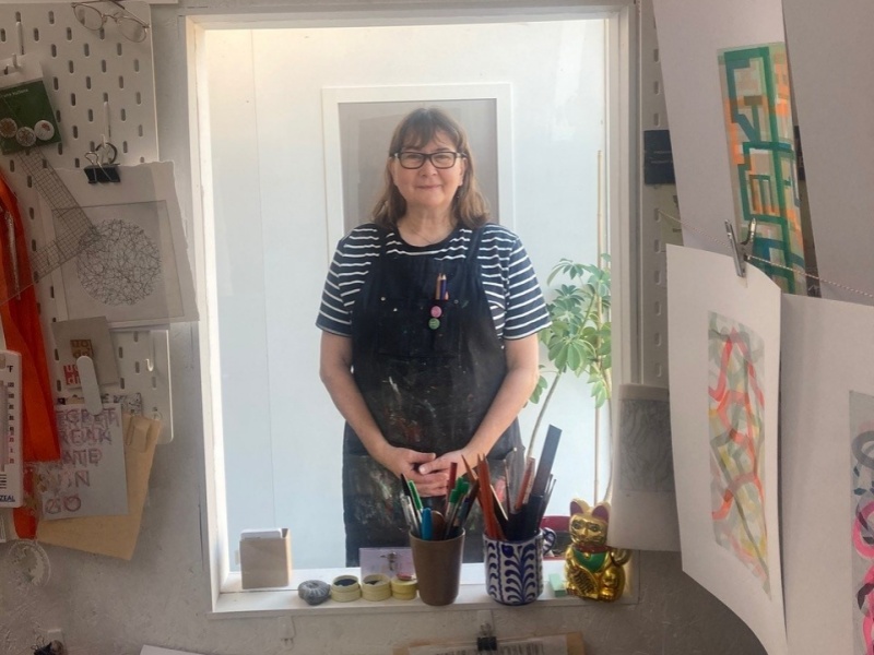 Emma stands behind an internal windowlike, hatch. She wears a blue and white short sleeved t-shirt, a navy blue apron and dark rimmed glasses. Her hands are clasped. In front of the hatch, on walls and table, are pieces of artwork, paper and equipment.