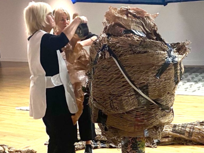 Disabled Artist Grace Currie stands in a gallery space and is wrapped in layers of packaging by two women. Grace's feet protrude from the bottom of the package. Her face, head, body and hands are all obscured. The performance was part of WAIWAV.