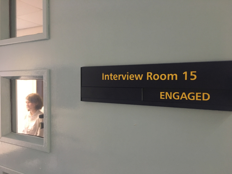 A film still of a grey wall with a large sign with the words 'Interview Room 15, Engaged' on it in yellow. To the left are two small windows and a woman is visible through the lower one. 