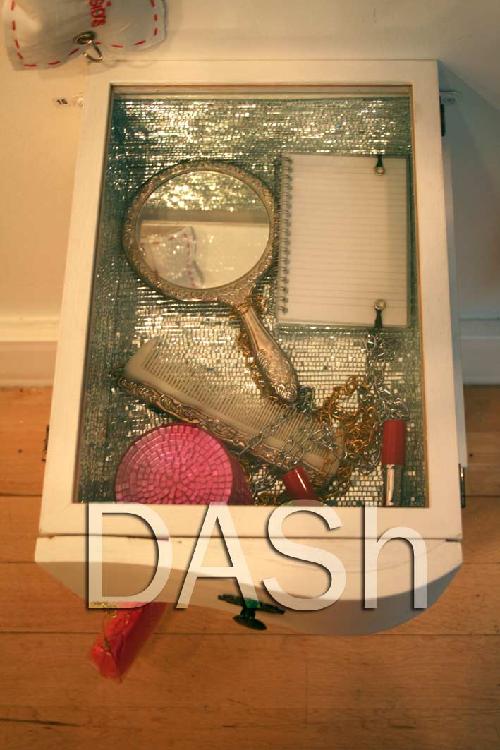 The image shows an artwork by Jenny Brown.  A glass lid shows the inside of a drawer.  The interior of the drawer is lined with tiny mirror tiles, a hand mirror, a notebook, a comb, and lipstick are on display in the drawer.  original artwork by Jenny Brown, photographed by Paula Dower.