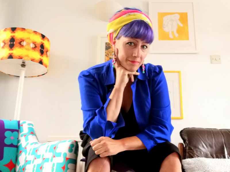 Kristina sits on a stool in a bright white room with retro accessories in orange and yellow. She wears a blue shirt, short black trousers and a scarf around her head. She rests her chin on her right hand.