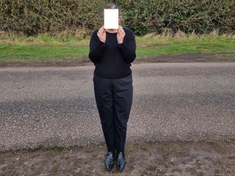 Mianam stands on a roadside, a green hedge behind. She wears black shoes, trousers and jumper and holds a piece of cream coloured card up to her face, hiding her from view.