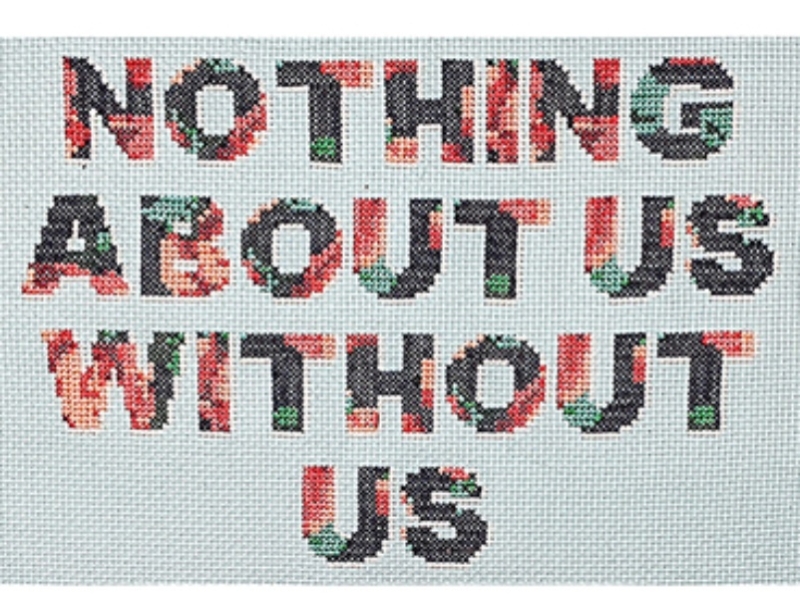 A cross-stitched panel. A white background with the words; Nothing About Us Without Us, stitched in bold capital lettering. The letters have a subtle floral pattern in navy blue, red and white vintage style.