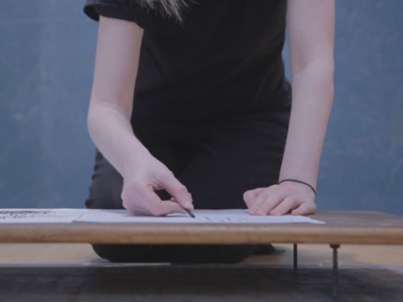 A cropped image showing the arms and hands of a person working on a raised wooden board at a table as they draw. They wear a black t-shirt with a black hair band around their left wrist and hold a piece of charcoal in their right hand.