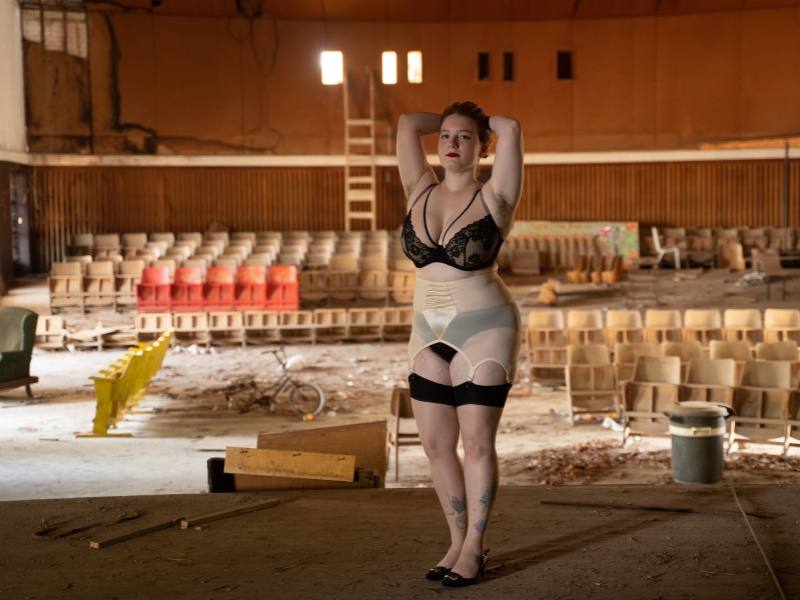 Stav stands on the stage of a run down, dilapidated theatre, arms up and hand resting on their head. They wear black underwear and stockings with a pale pink suspender belt.