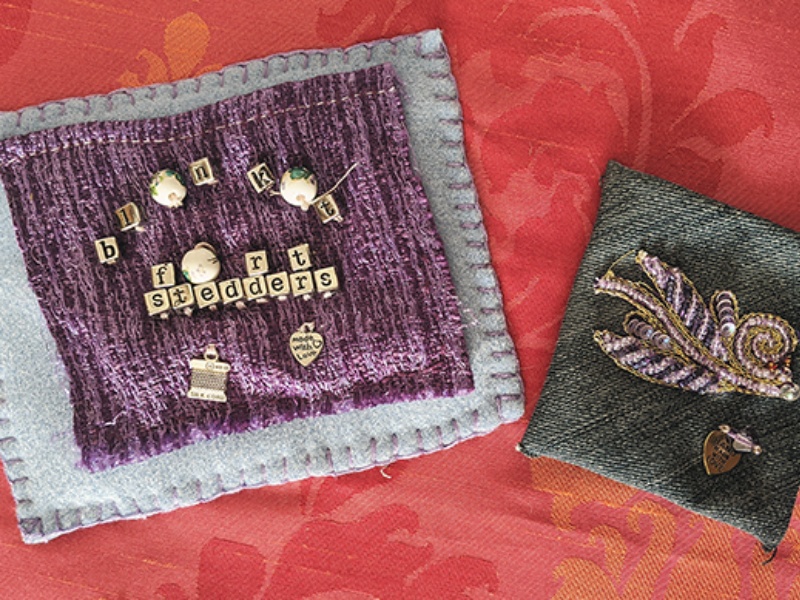 A pale blue square with smaller purple one stitched above. There are small square lettered beads attached spelling out 'blanket fort stedders.' Besides this, a small rectangular piece of denim with an embroidered leaf-like patch in gold embroidery.