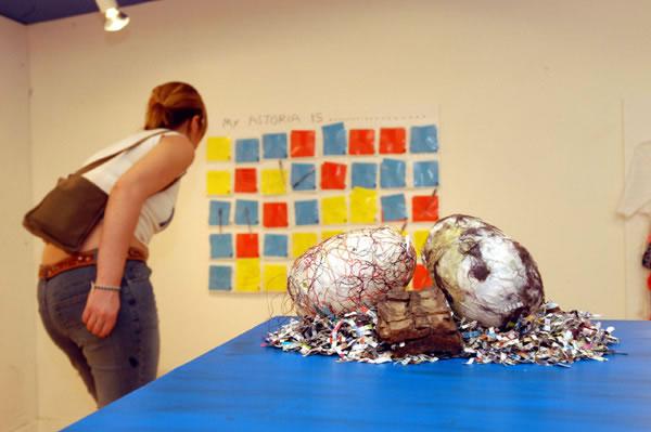 Three Astorian dinosaur eggs, the eggs are made of paper and textiles, they sit on top of shredded paper. Behind the eggs a gallery visitor is looking at artwork on the wall, photo by Anthea Bevan