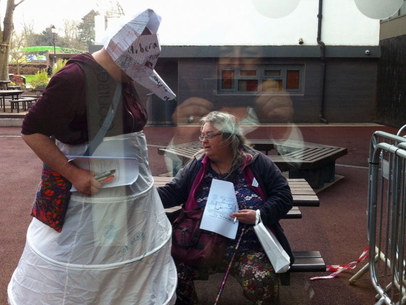 gobscure standing outdoors wearing a structured white petticoat which has been written on. The artist wears a 3D paper head mask with a large pointed nose that obscures their face. A seated woman appears to adjust the fabric.